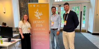 Ein Gruppenfoto von drei Personen, die neben einem großen, orange-rosa Farbverlaufs-Banner in einem Foyer stehen. Das Banner trägt in großen Lettern die Aufschrift „DESRIST 2026“ und bewirbt die „21st International Conference on Design Science Research in Information Systems and Technology“ vom 8. bis 10. Juni 2026 in Münster. Links neben dem Banner steht eine Frau mit langem, hellem Haar, bekleidet mit einem weißen Kurzarm-Oberteil mit Lochstickerei und einem dunklen Rock. Rechts vom Banner stehen zwei Männer: Der linke trägt eine Brille, ein hellblaues Hemd, Jeans und einen braunen Gürtel; der rechte ist der Mann aus dem ersten Bild, gekleidet in einen dunkelblauen Pullover über einem weißen Hemd und helle Hosen. Alle drei tragen Konferenz-Namensschilder um den Hals und blicken lächelnd in die Kamera. Im Hintergrund sind verglaste Doppeltüren zu sehen, die nach draußen ins Grüne führen, sowie ein Tisch mit einem Monitor am linken Bildrand.
